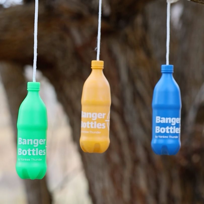 Three shatterproof Banger Bottles Targets by Yankee Thunder hang from strings outdoors. The bottles come in green, yellow, and blue, each featuring 