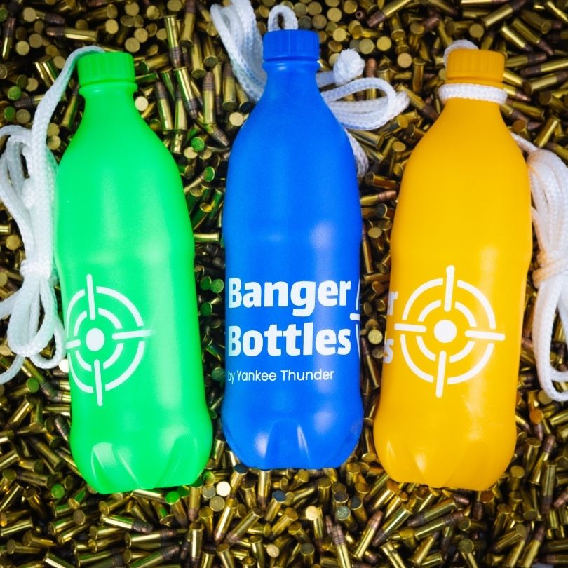 Three shatterproof Banger Bottles Targets by Yankee Thunder, in green, blue, and yellow, rest on a bed of brass bullet casings. Each durable and reusable bottle has a white cap and an attached white rope. The bottles sport an easy-to-see target logo design.