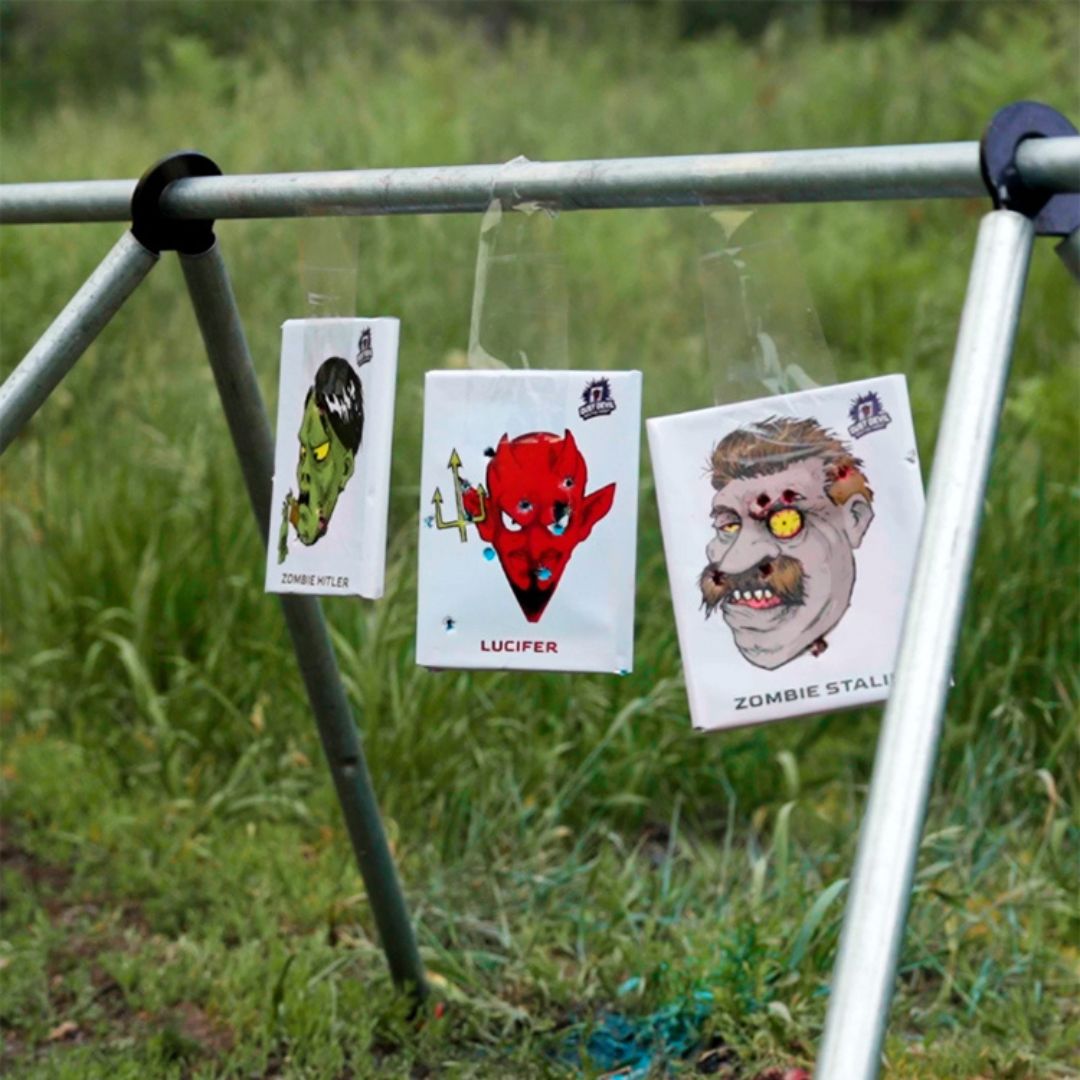 Three illustrated shooting targets from the Dust Devil - Special Donkey Edition by Yankee Thunder dangle from a metal frame in a grassy field, made from colored chalk for immediate feedback. The targets depict caricatures labeled 