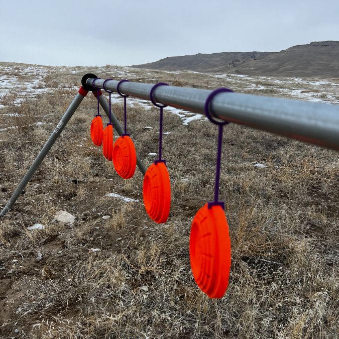 A row of five bright orange clay shooting targets from the Yankee Thunder Bang Bang Accessory Kit hangs from purple cords attached to a silver metal bar equipped with AR500 steel gongs. The setup is situated in an open, grassy field with some snow patches and distant hills visible under a cloudy sky.