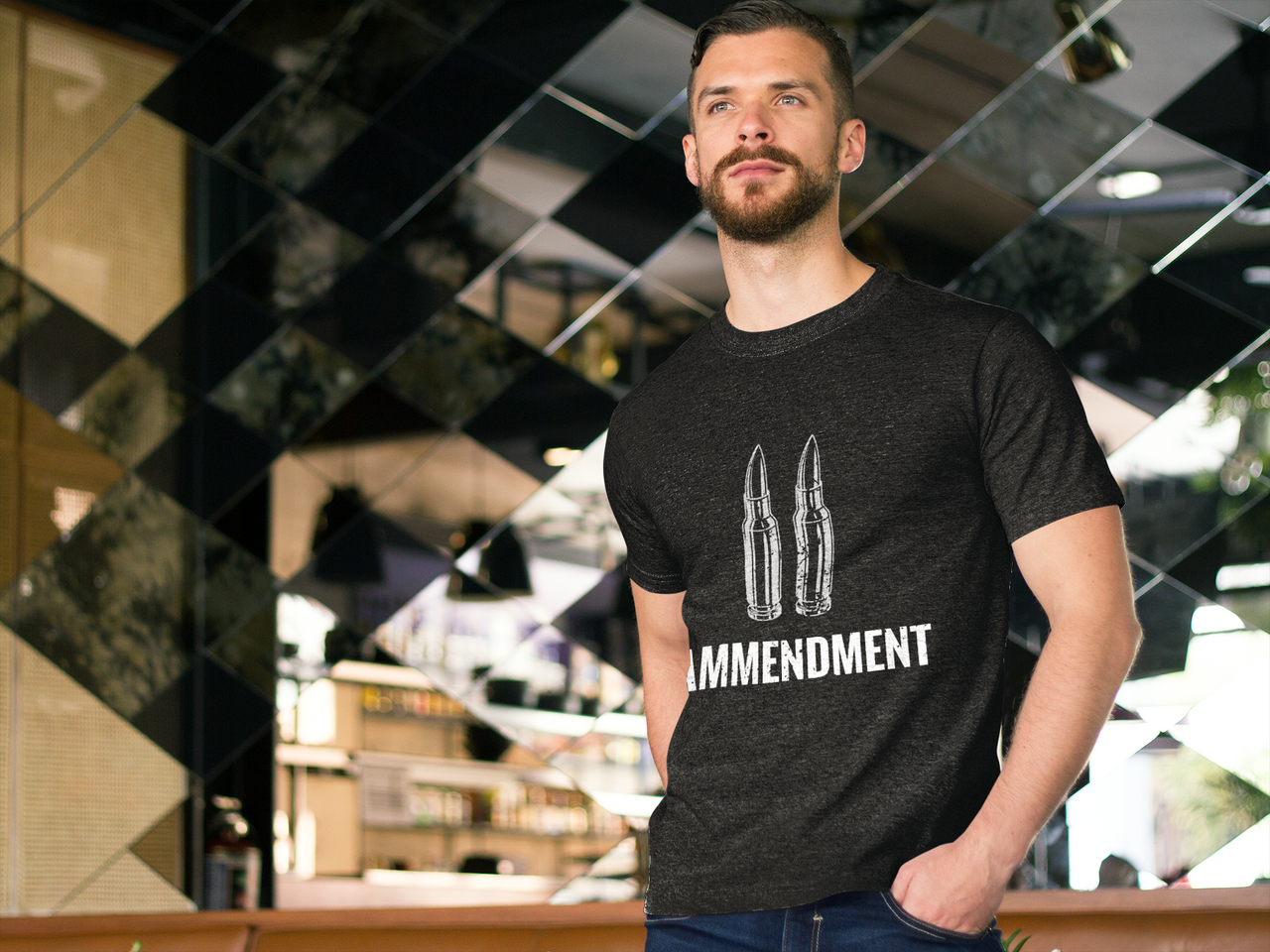 A man with short hair and a beard stands indoors in front of a reflective, patterned wall. He is dressed in a dark Yankee Thunder 2nd Amendment Tee, which features two bullet illustrations above the words "2nd Amendment," subtly emphasizing the message to protect our freedom. With his hands in his pockets, he gazes off to the side.